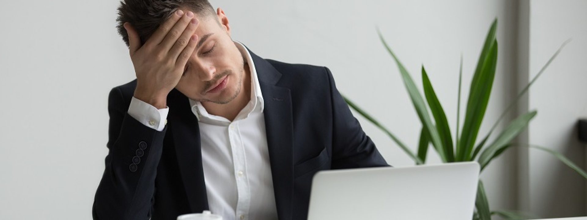 Frustrated fatigued millennial businessman having strong headache tired from laptop use touching forehead, stressed overworked company ceo suffers from migraine at work, aching head in office concept